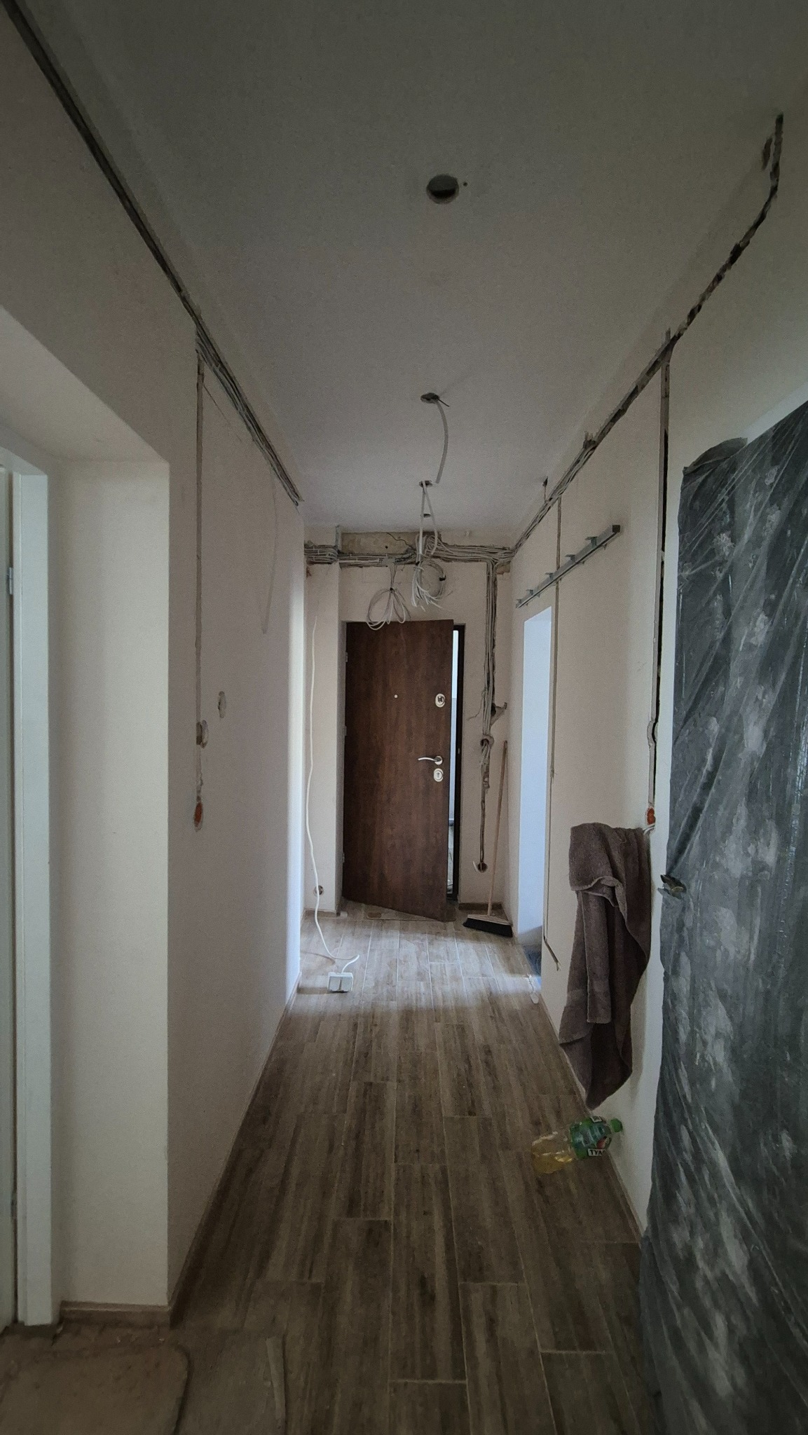 Korytarz w trakcie remontu z widocznymi przewodami elektrycznymi na ścianach i suficie. Częściowo zabezpieczone ściany folią malarską. Widoczne kable do oświetlenia.