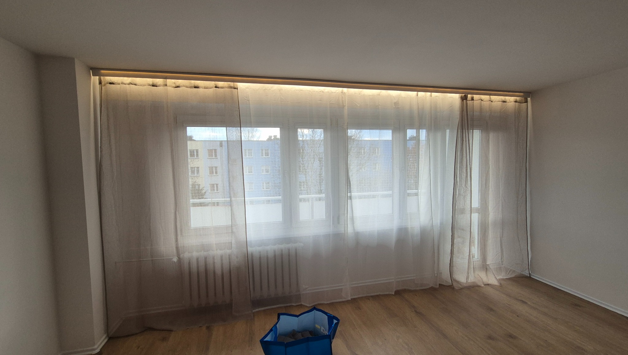 Jasne, zwiewne zasłony z podświetleniem LED nad oknem w pokoju z parkietem. Widok na budynek zza okna. Niebieski kosz na podłodze dodaje koloru.
