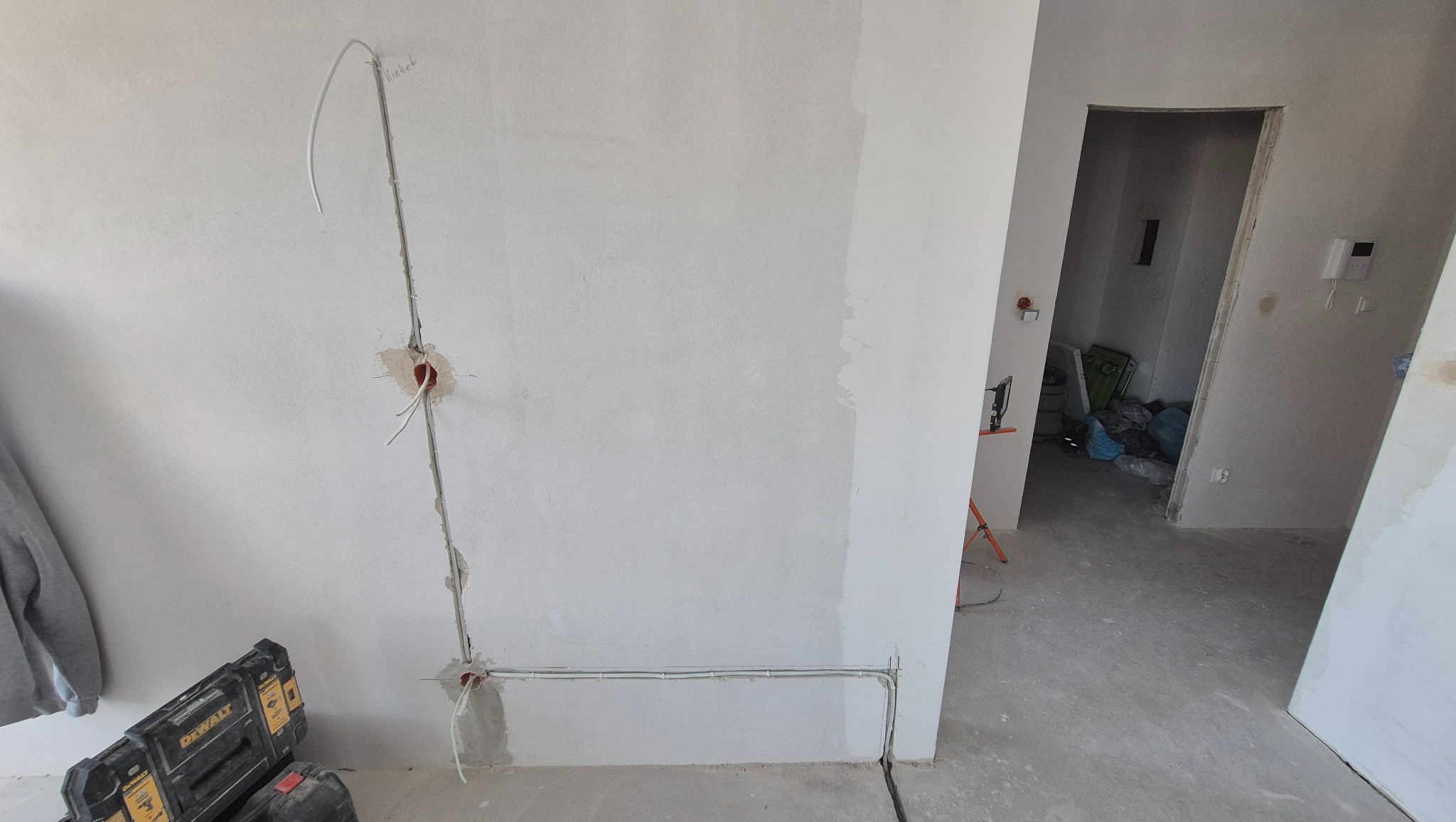 Ściana w trakcie remontu z poprowadzonymi przewodami elektrycznymi w peszlu i puszkami pod gniazdka. Widoczny sprzęt DeWalt i otwarte przejście do innego pomieszczenia.
