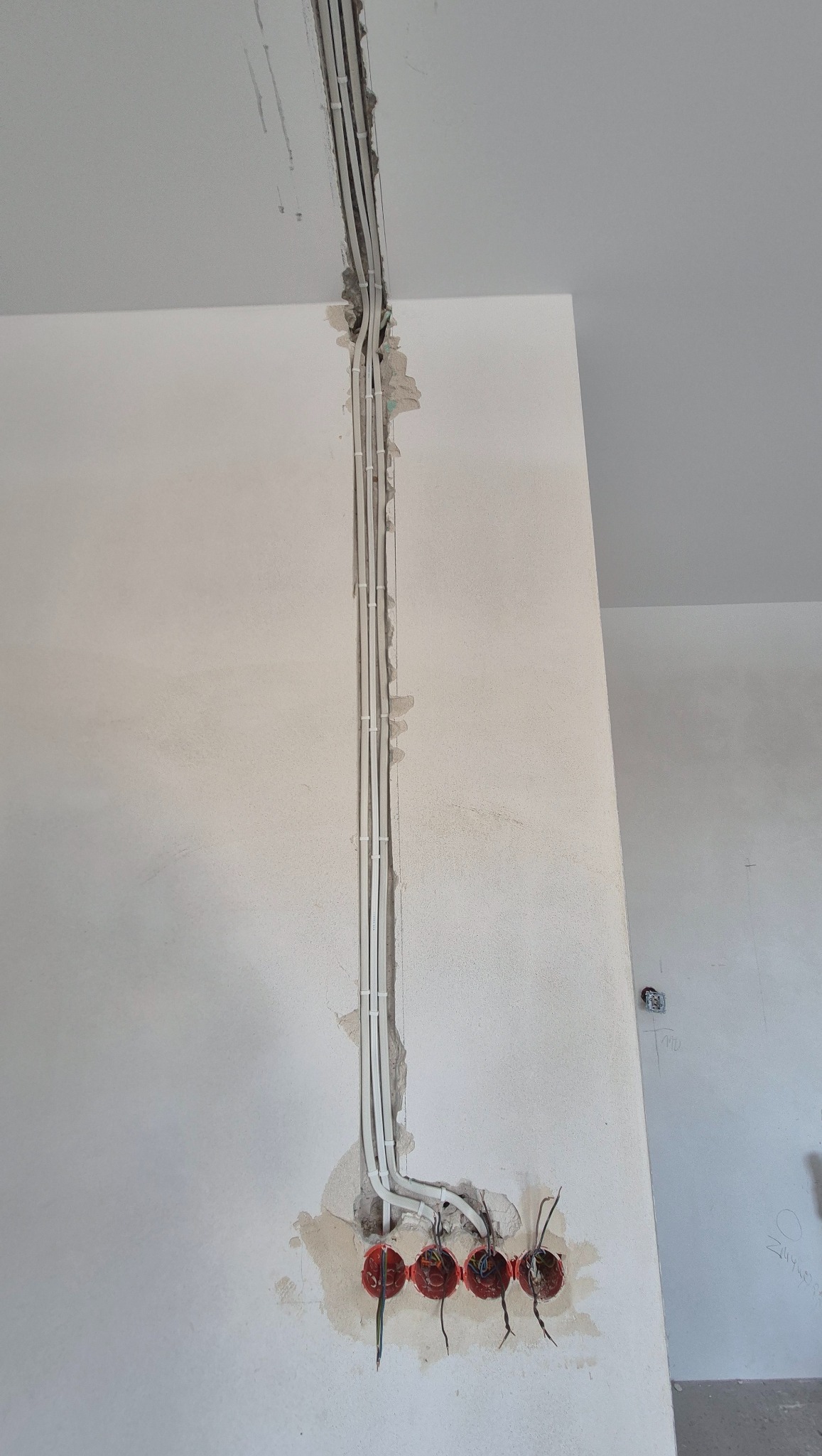 Białe kable elektryczne w bruzdach ściany, prowadzące do czerwonych puszek instalacyjnych. Widoczne niedoskonałości tynku wokół instalacji. Ściana w surowym stanie.