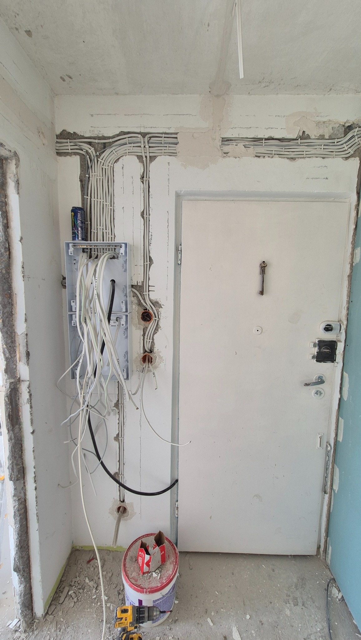 Rozdzielnia elektryczna z przewodami w surowym wnętrzu, widoczne puszki instalacyjne, puszka z napojem na rozdzielni, narzędzia na podłodze. Prace elektryczne w trakcie remontu.