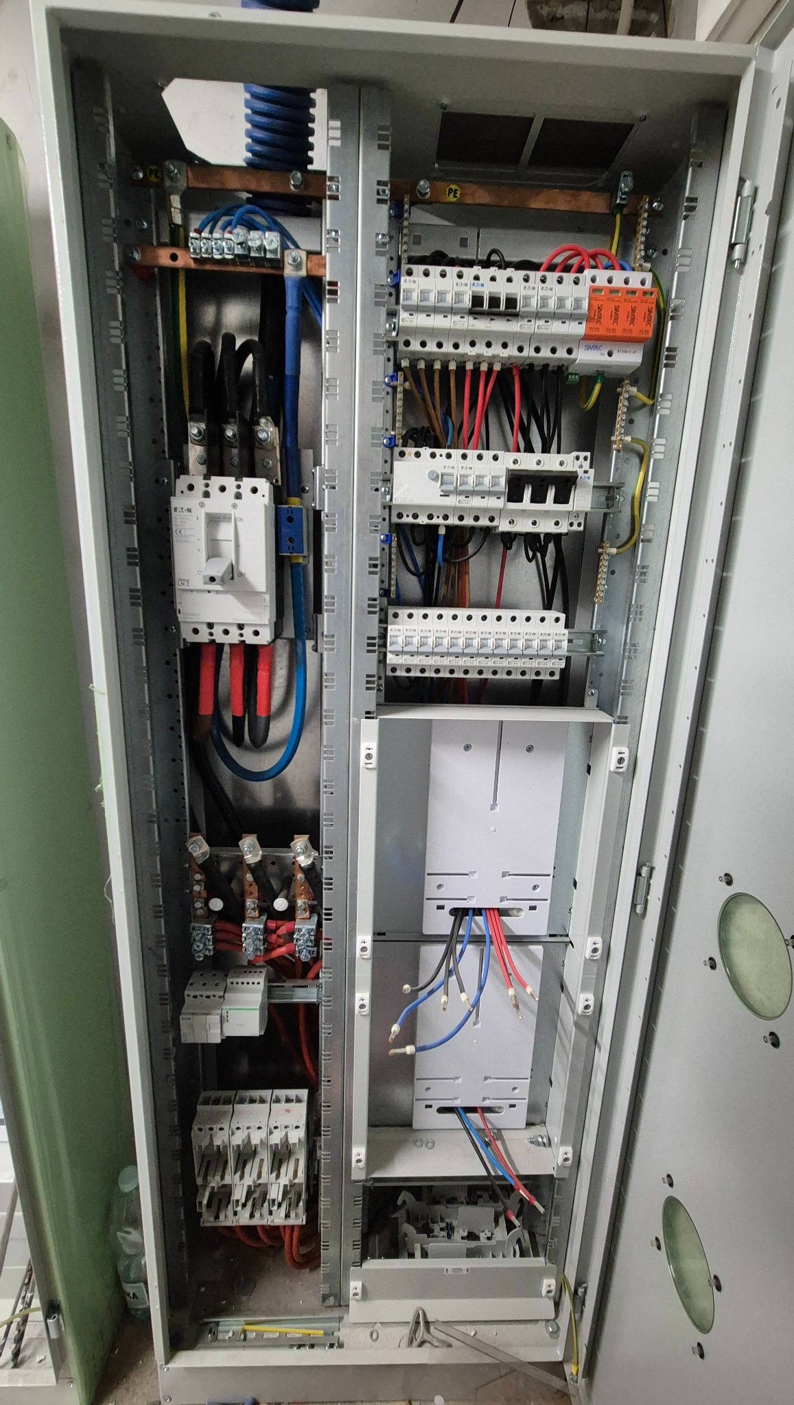 Otwarta szafa elektryczna z widocznymi bezpiecznikami, przewodami i elementami instalacji. Metalowa konstrukcja, różnokolorowe kable. Widok z bliska na wnętrze rozdzielnicy.