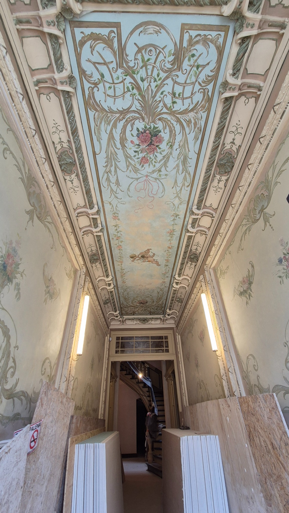 Korytarz z dekoracyjnymi malowidłami na suficie i ścianach w stylu retro. Widoczne panele i materiały budowlane, sugerujące prace remontowe w toku.