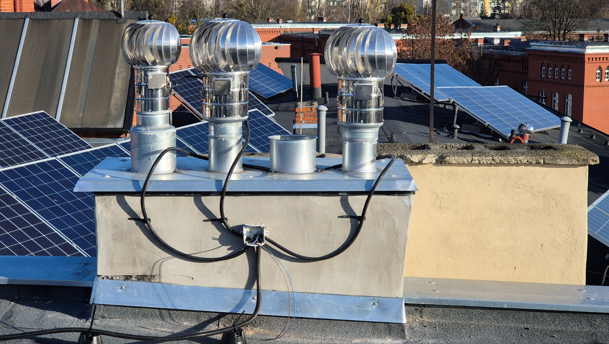 Instalacja turbin wentylacyjnych i paneli słonecznych na dachu budynku. Widoczne przewody elektryczne i puszka przyłączeniowa. Ujęcie z bliska, w słoneczny dzień.