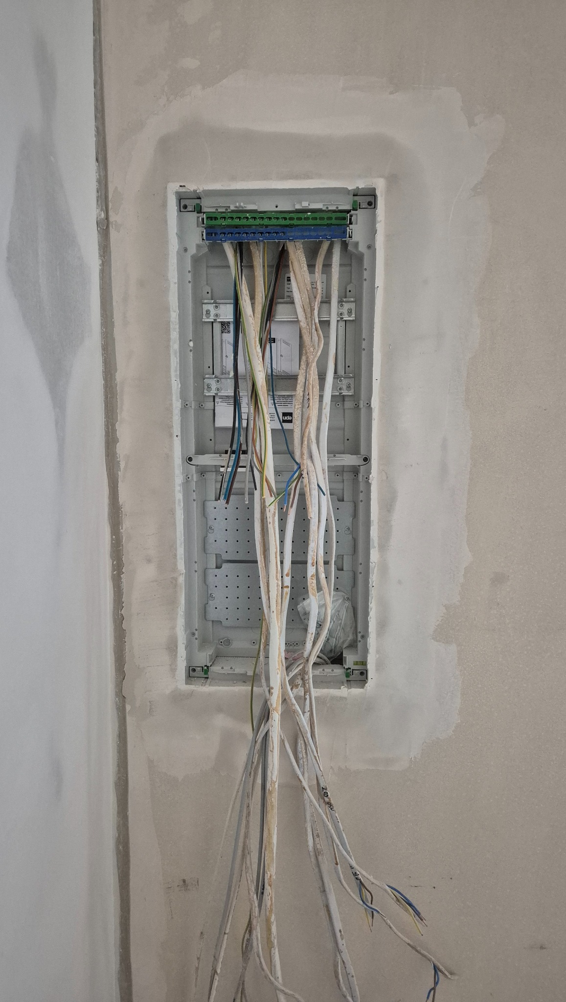 Wnęka w ścianie z zamontowaną skrzynką elektryczną i wystającymi przewodami w trakcie prac instalacyjnych. Widoczne złącza i kable w różnych kolorach.