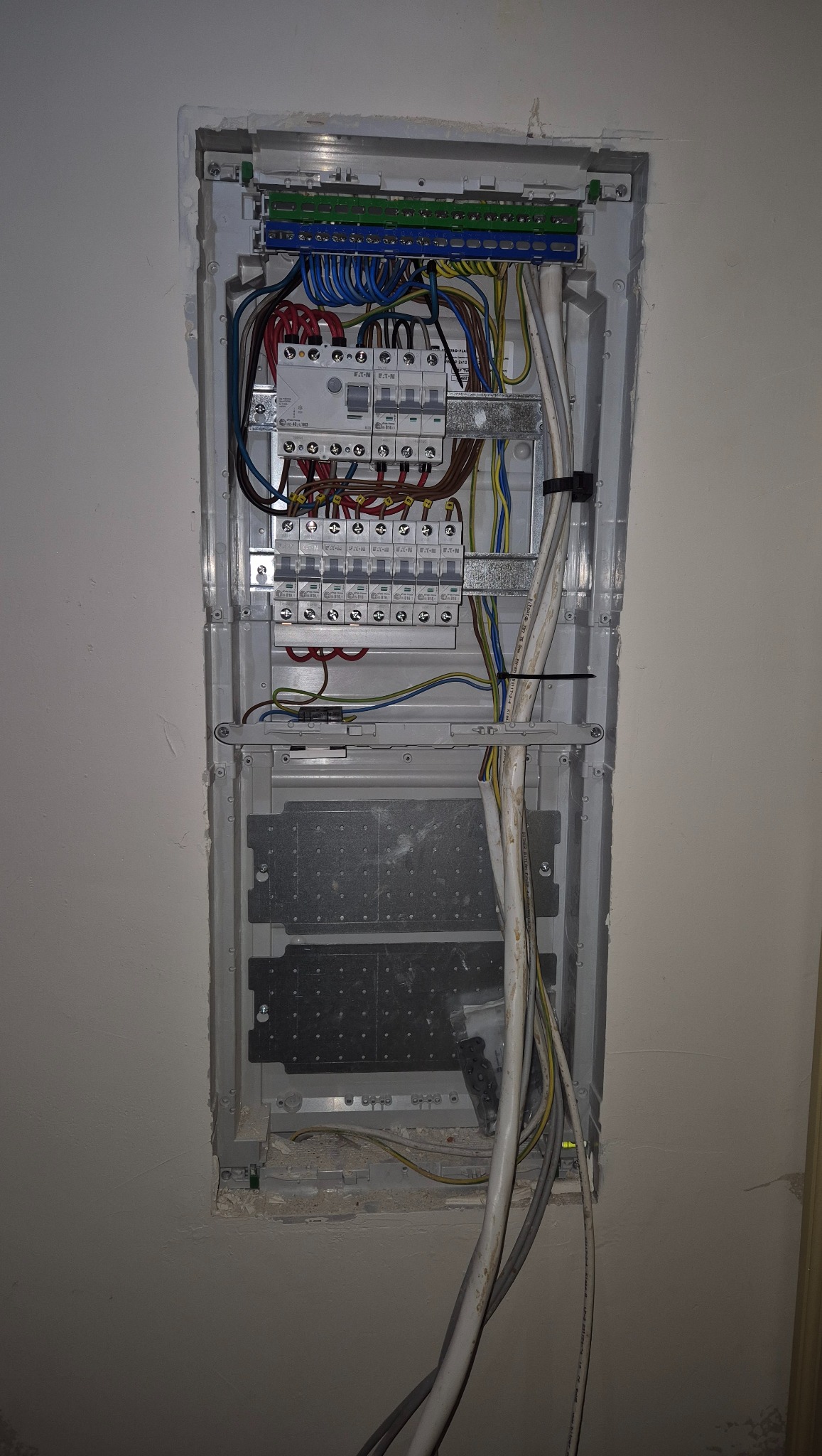 Rozdzielnica elektryczna wnękowa z widocznymi przewodami, bezpiecznikami i listwami zaciskowymi. Instalacja w trakcie montażu, widoczne niedoskonałości wykończenia ściany.