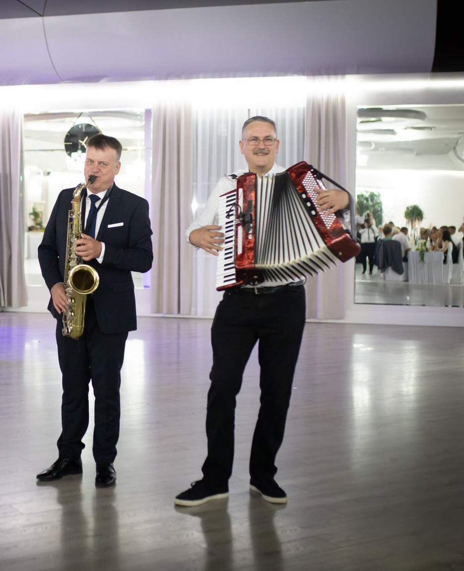 Duet muzyczny: saksofonista w garniturze i akordeonista w koszuli, występują na jasnej sali balowej z lustrami i gośćmi w tle. Profesjonalne oświetlenie.