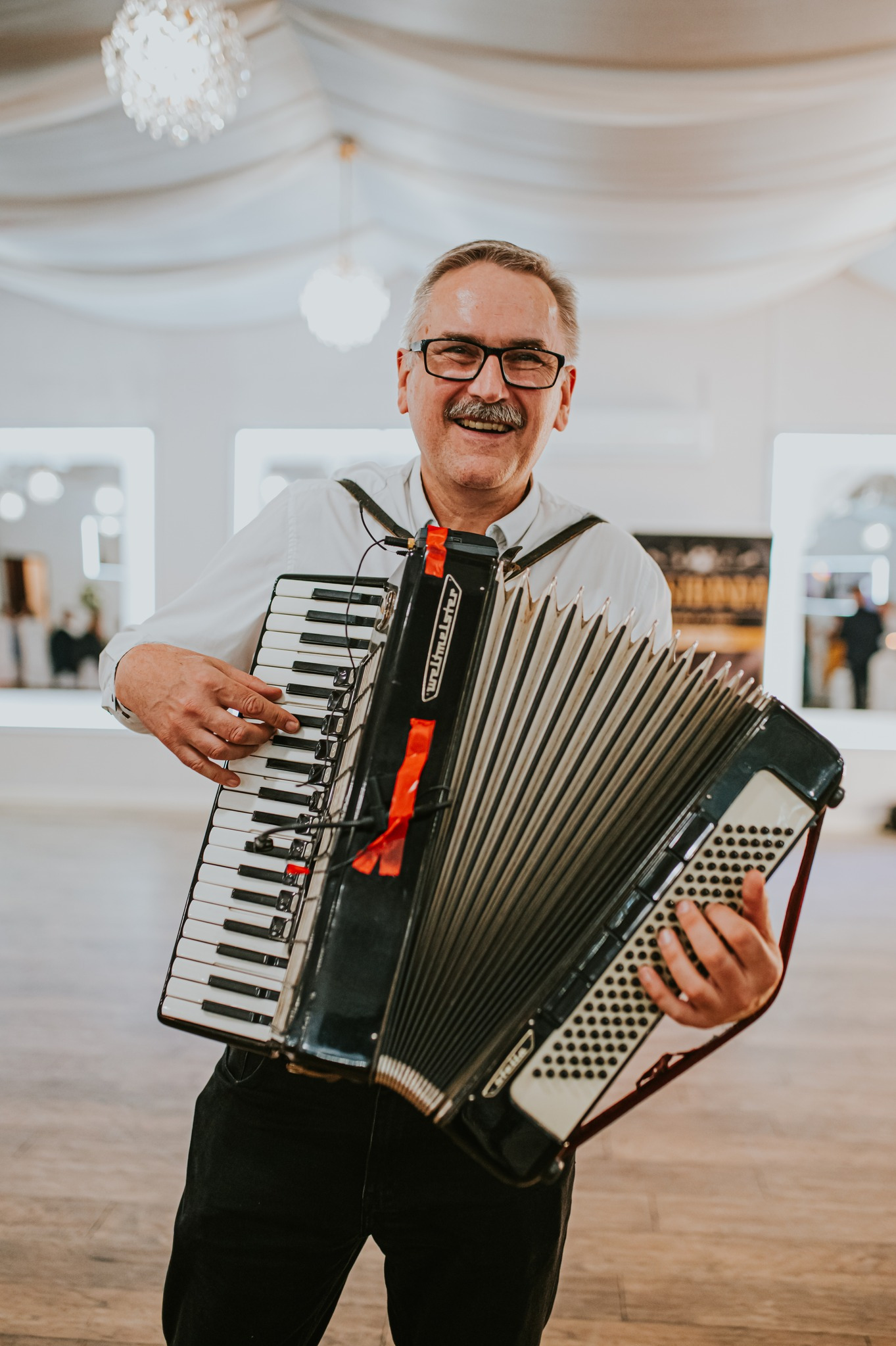 Uśmiechnięty akordeonista w okularach, grający na czarno-białym akordeonie Weltmeister z czerwoną wstążką, w jasnym wnętrzu sali.