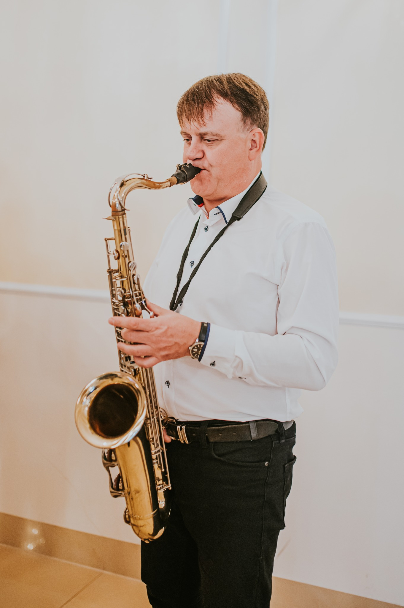 Saksofonista w białej koszuli gra na saksofonie tenorowym. Widoczny zegarek na nadgarstku i czarny pasek od saksofonu. Jasne tło.