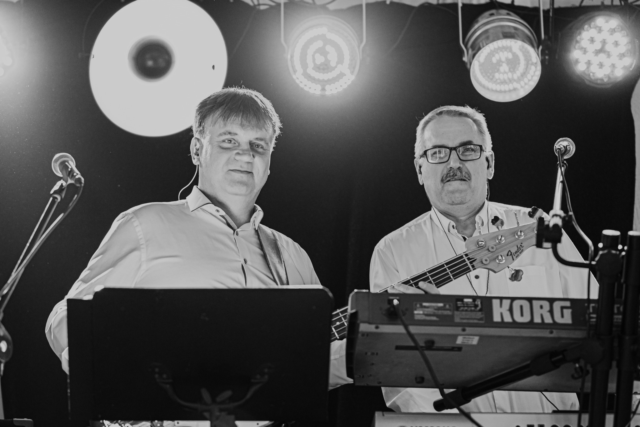 Czarno-białe zdjęcie dwóch muzyków z instrumentami (gitara basowa Fender, keyboard Korg) na scenie, oświetlonych reflektorami. Widoczne mikrofony i monitory odsłuchowe.