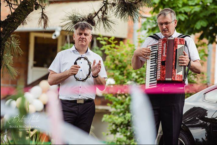Dwóch muzyków w plenerze: jeden gra na akordeonie, drugi trzyma tamburyn. W tle budynek i auto, w pierwszym planie dekoracje z balonów. Ujęcie z dołu.