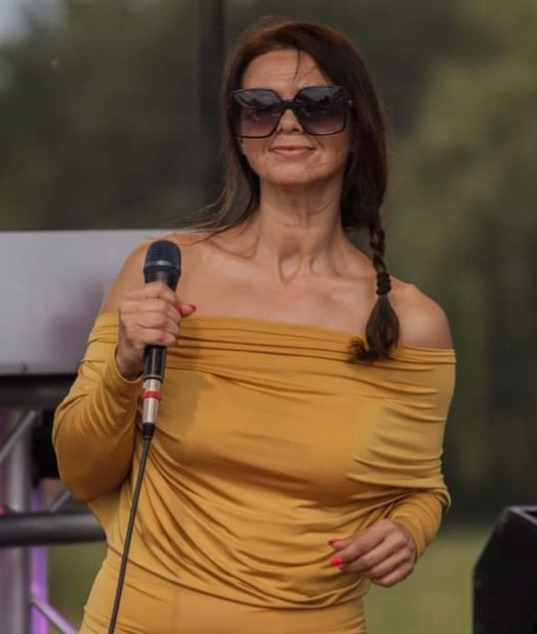 Kobieta w musztardowej sukience śpiewa do mikrofonu na tle zieleni. Uśmiechnięta piosenkarka w okularach przeciwsłonecznych, z warkoczem, podczas występu na scenie.