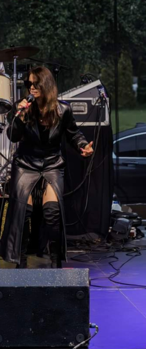 Wokalistka w czarnym, skórzanym płaszczu i okularach śpiewa do mikrofonu na scenie podczas koncertu plenerowego. W tle perkusja i sprzęt nagłośnieniowy.