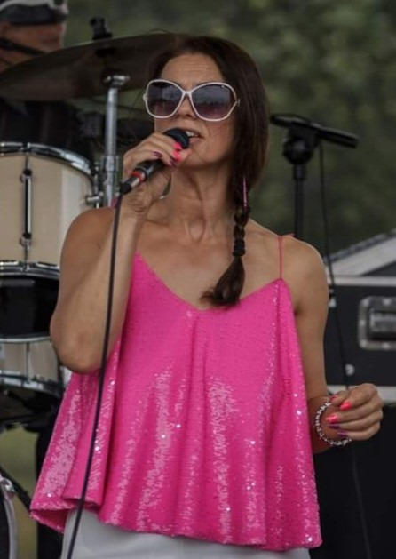 Wokalistka w różowej cekinowej bluzce śpiewa do mikrofonu na scenie, w tle perkusja i sprzęt muzyczny. Ujęcie z występu zespołu muzycznego.