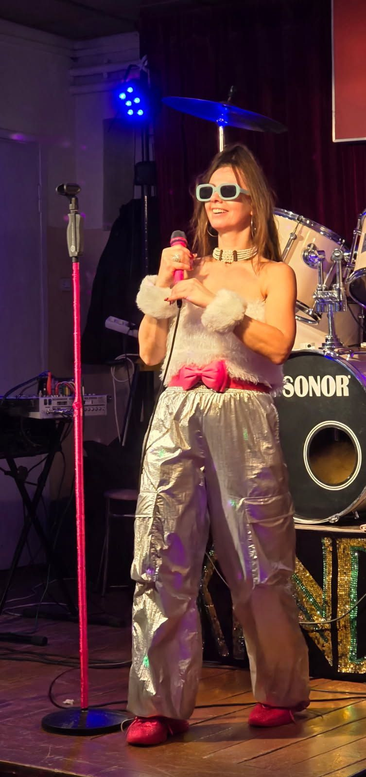 Wokalistka w srebrnych spodniach i różowym mikrofonem na scenie z perkusją Sonor w tle, oświetlona niebieskim światłem. Nietypowy strój i okulary.