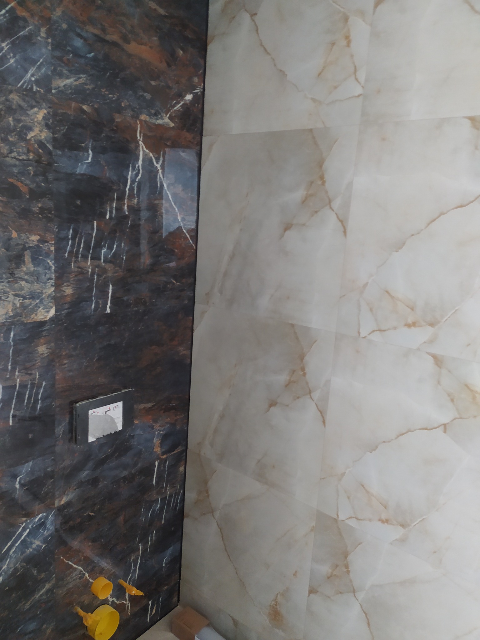 Ułożone płytki imitujące marmur w odcieniach bieli i brązu, kontrastujące z ciemnymi płytkami z widocznym osprzętem. Widoczne narzędzia glazurnicze.