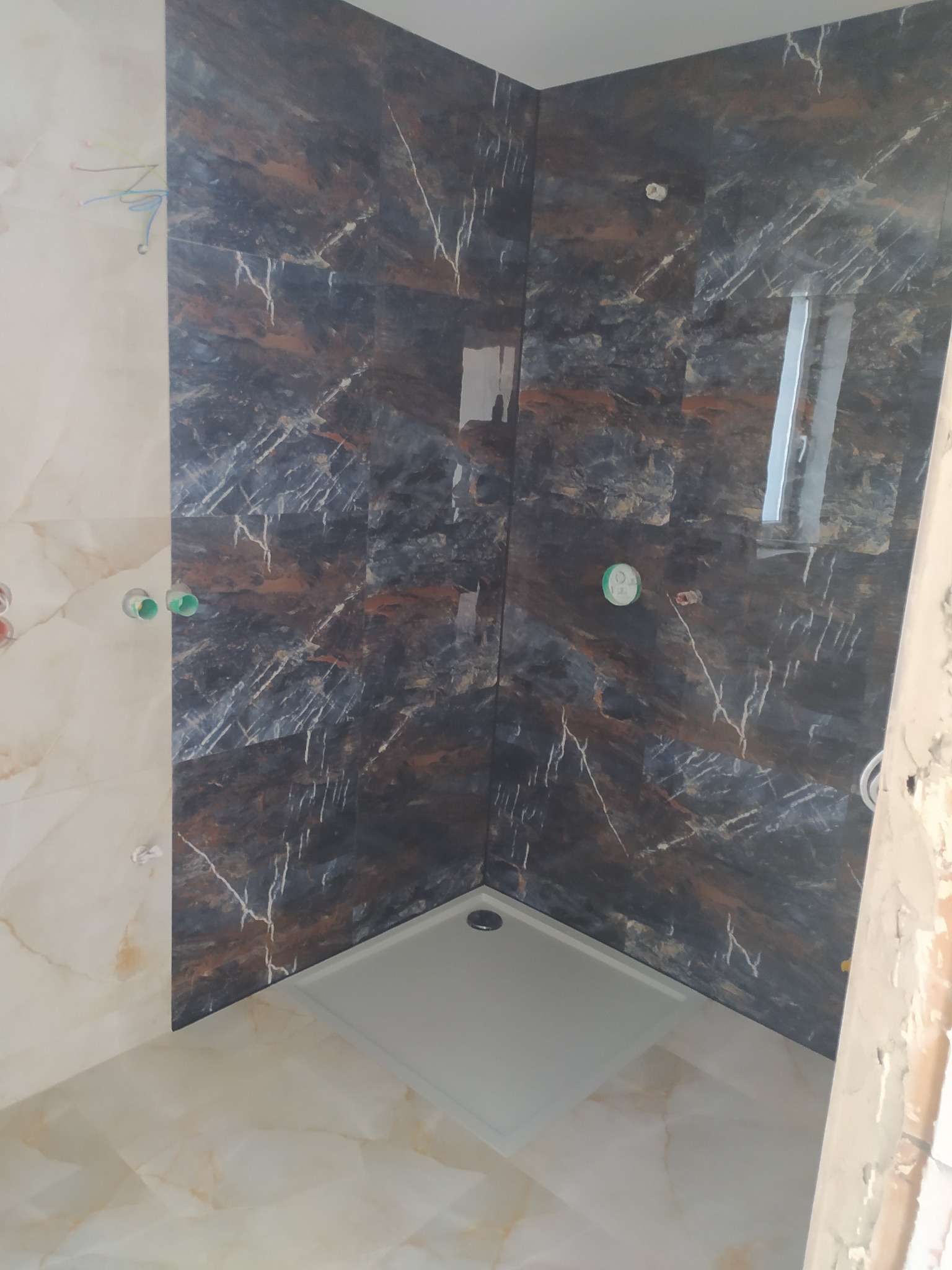 Wykończona kabina prysznicowa z ciemnymi, marmurkowymi płytkami i brodzikiem. Widoczne elementy instalacji wodnej. Ukończona realizacja w łazience.