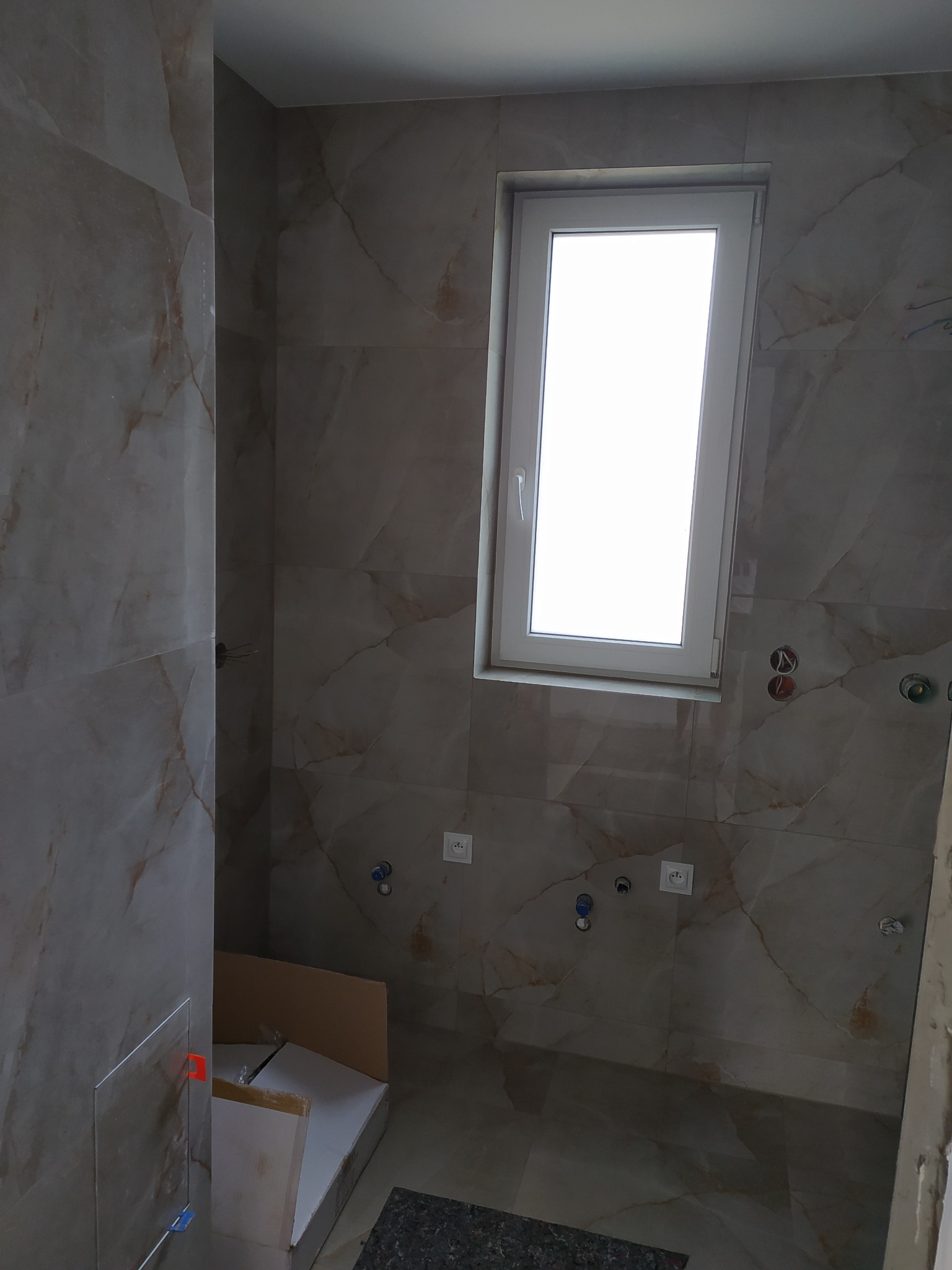 Wykończona łazienka z beżowymi płytkami imitującymi marmur, białe okno, widoczne przyłącza wodne i elektryczne, karton z materiałami budowlanymi w rogu.