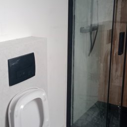 Solid-bud - Nowoczesna łazienka: podwieszana toaleta z czarnym przyciskiem spłukującym i kabina prysznicowa z ciemną ramą. Minimalistyczny design, jasne ściany.