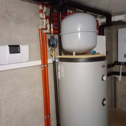 Solid-bud - Szary bojler z podłączeniami rur miedzianych i czarnych, zamontowany w pomieszczeniu z szarą płytką na ścianie i podłodze. Widoczne elementy instalacji grzewczej.