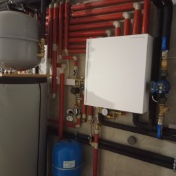 Solid-bud - Rozbudowana instalacja grzewcza z licznymi rurami (czerwone, czarne) i białym modułem DEPRO na ścianie z płytek. Widoczny zbiornik i niebieskie naczynie przeponowe.