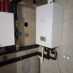 Solid-bud - Biały piec gazowy dwufunkcyjny zamontowany na szarej, kafelkowanej ścianie. Widoczne rury, zawory i instalacja. Nowoczesne wykończenie łazienki lub kotłowni.