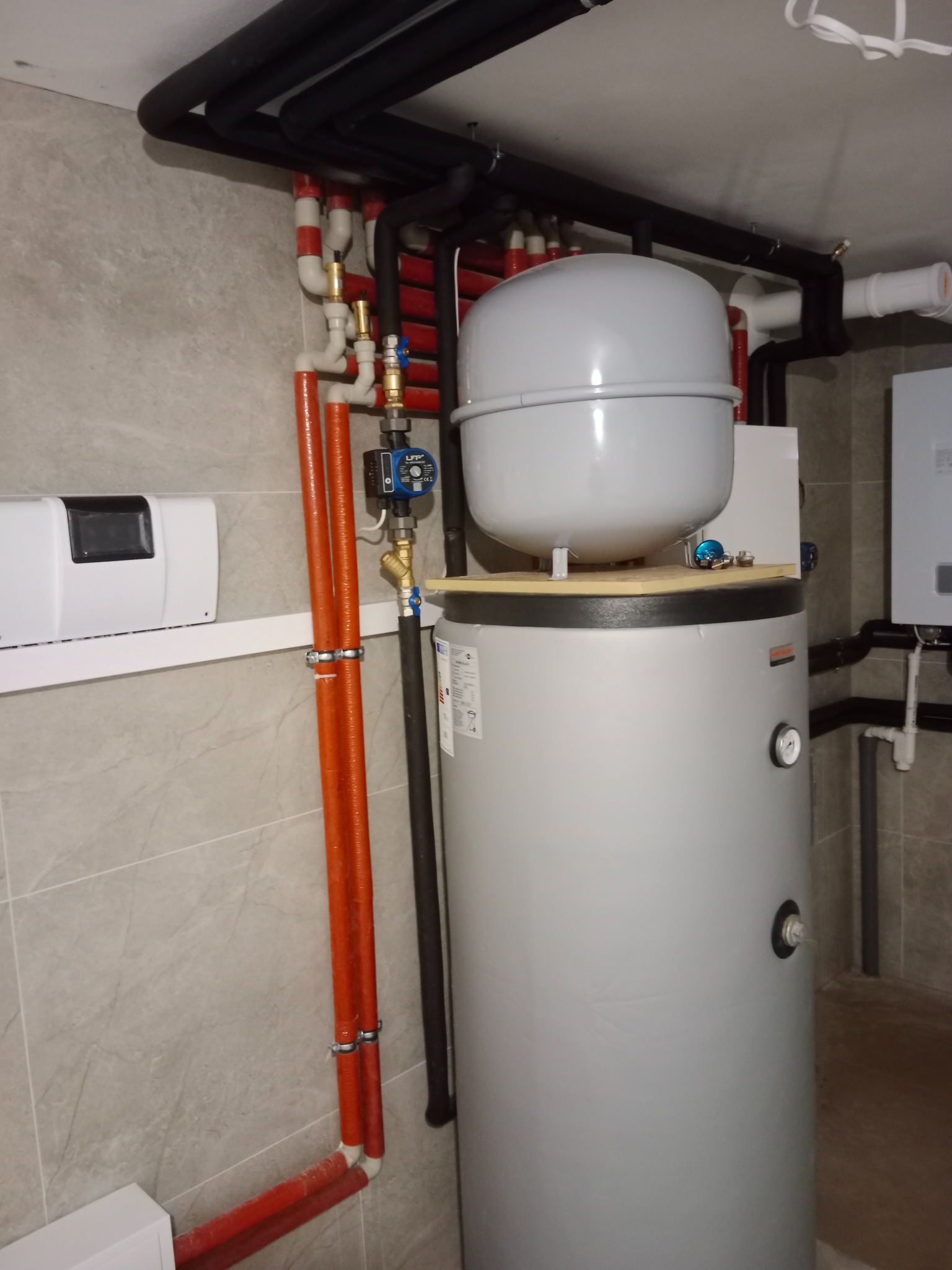 Szary bojler z podłączeniami rur miedzianych i czarnych, zamontowany w pomieszczeniu z szarą płytką na ścianie i podłodze. Widoczne elementy instalacji grzewczej.