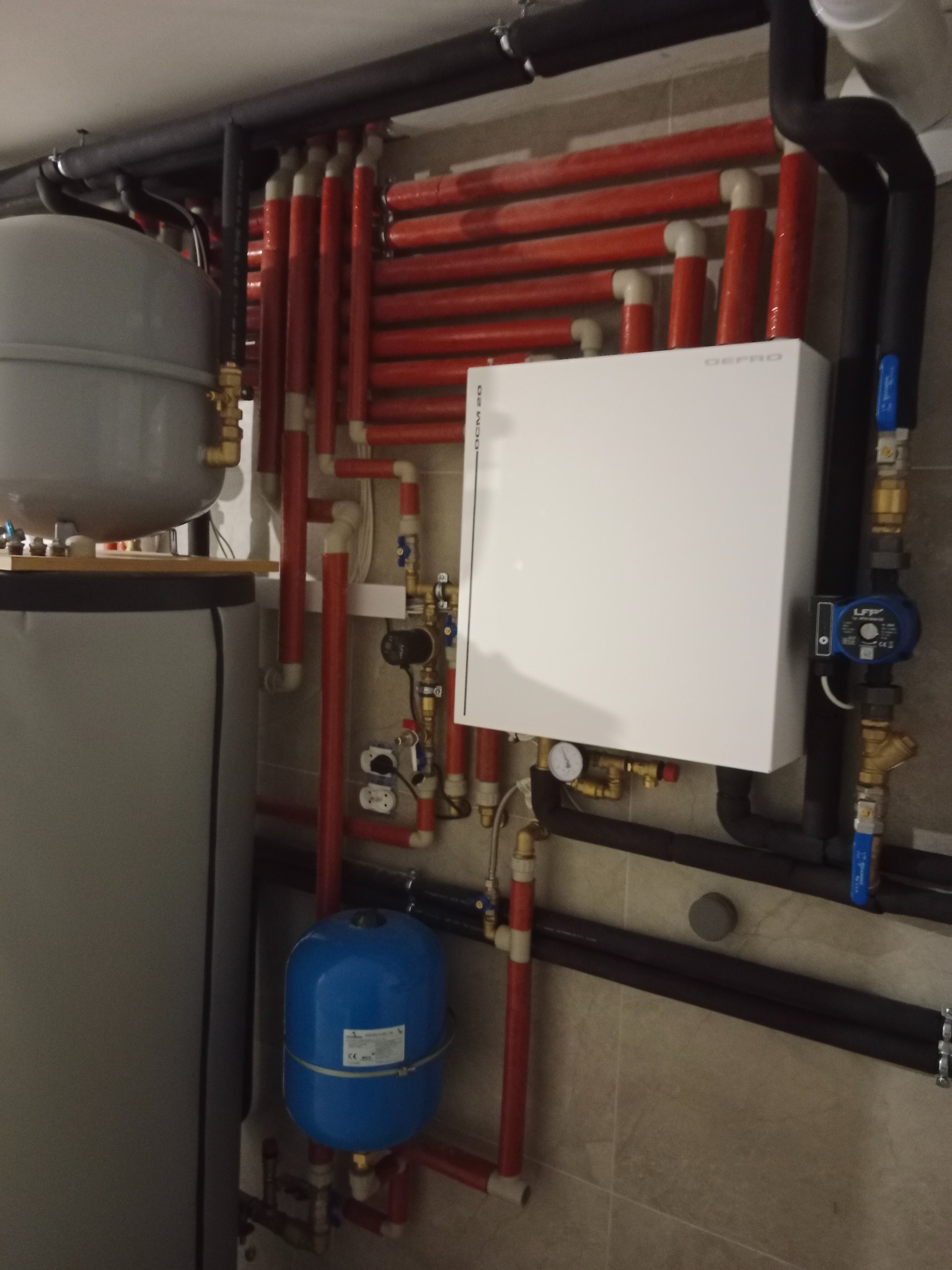 Rozbudowana instalacja grzewcza z licznymi rurami (czerwone, czarne) i białym modułem DEPRO na ścianie z płytek. Widoczny zbiornik i niebieskie naczynie przeponowe.