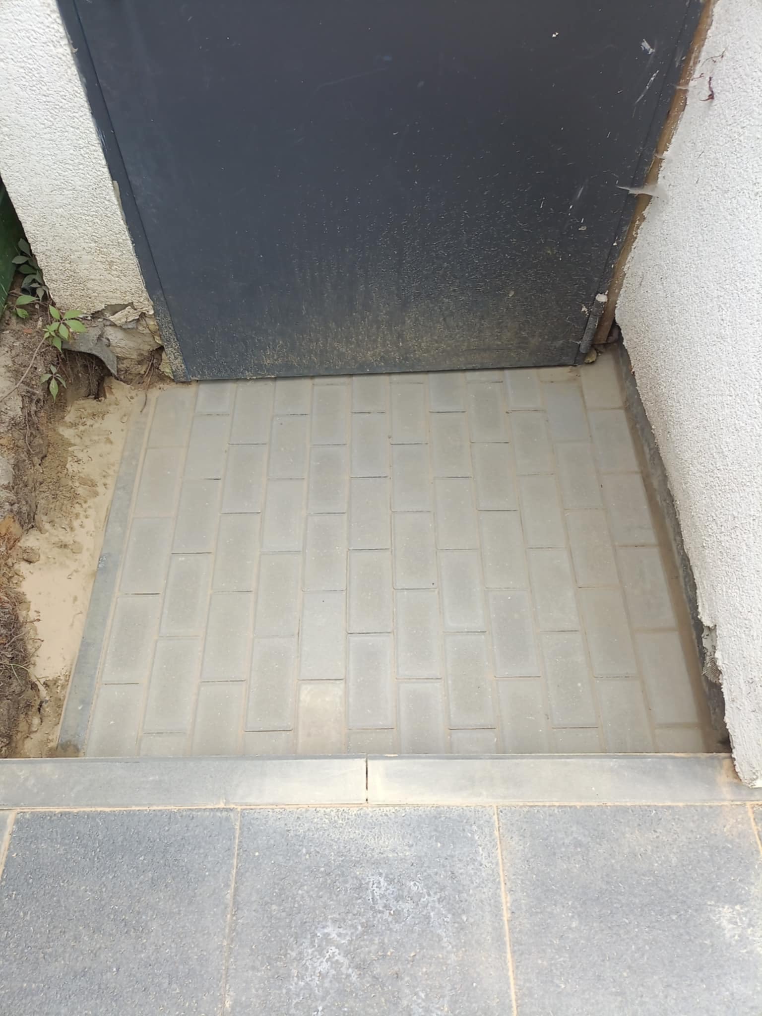 Ułożona szara kostka brukowa przed ciemnymi drzwiami wejściowymi, widok z góry. Krawężnik oddziela kostkę od ciemniejszej nawierzchni.