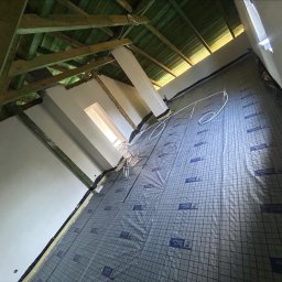 Wiktor Zarzycki - Skośny kadr na poddasze w trakcie izolacji. Widoczne drewniane elementy konstrukcyjne, okno dachowe i folia izolacyjna z nadrukiem siatki i logo 'MONTER Quality'.