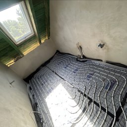 Wiktor Zarzycki - Białe rury ogrzewania podłogowego ułożone na szarej macie w surowym wnętrzu z oknem dachowym i widocznymi elementami instalacji na ścianie.