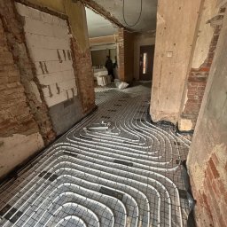 Wiktor Zarzycki - Białe rury ogrzewania podłogowego ułożone na czarnej folii w remontowanym wnętrzu z odsłoniętą cegłą i widoczną instalacją elektryczną.