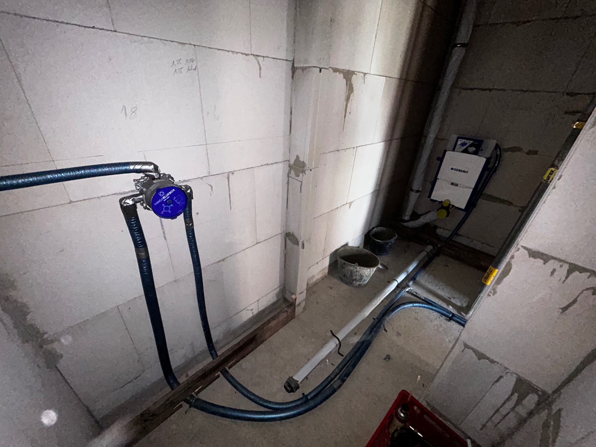 Surowy stan: Instalacja wodna z niebieskim licznikiem Hansa Bluebox na tle ściany z betonu komórkowego. Widoczne rury i elementy instalacji podtynkowej Geberit.