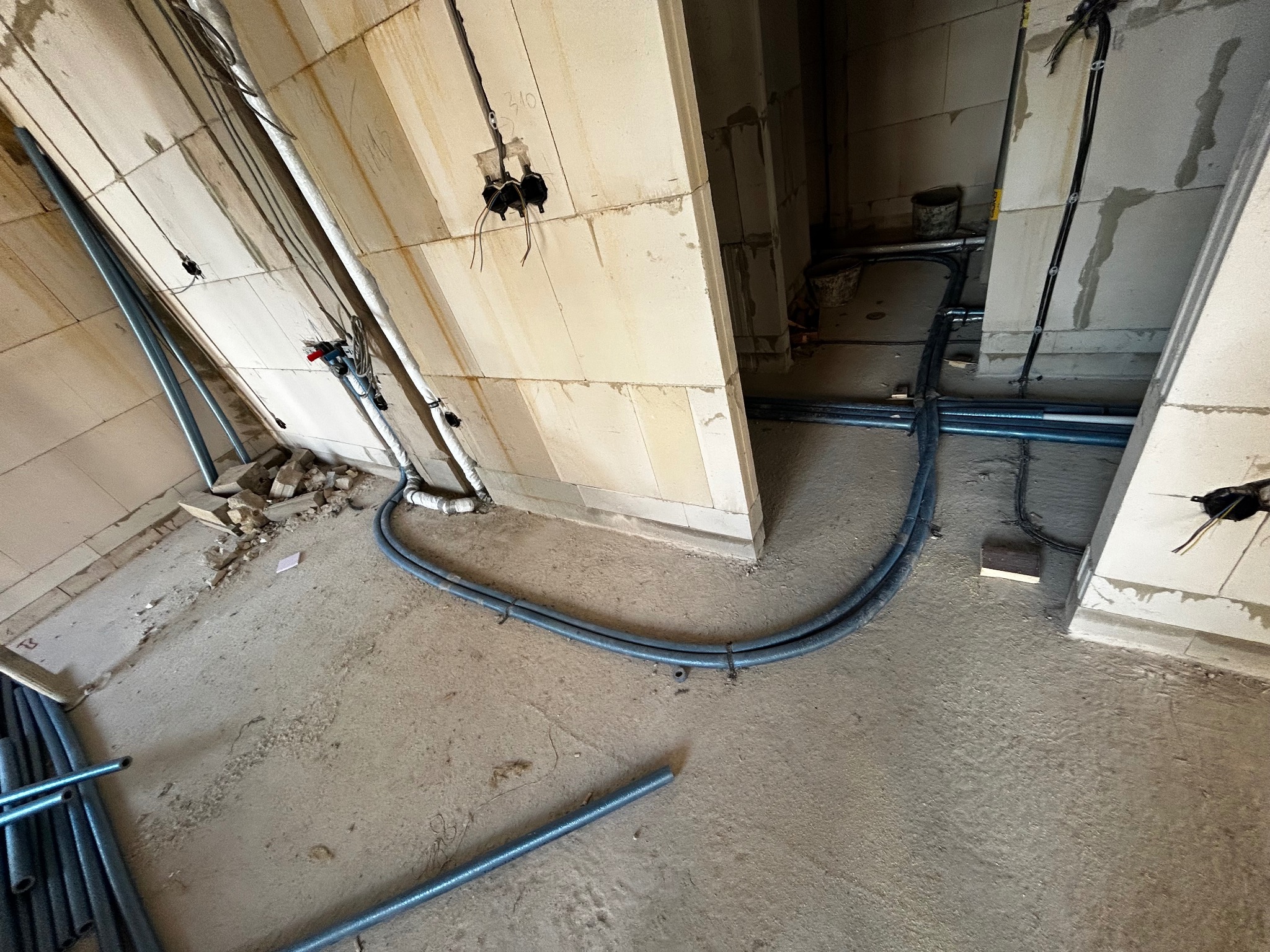 Szare rury instalacji ogrzewania podłogowego ułożone na surowym betonie w trakcie montażu w nowym budynku z białych bloczków. Widoczne kable elektryczne.