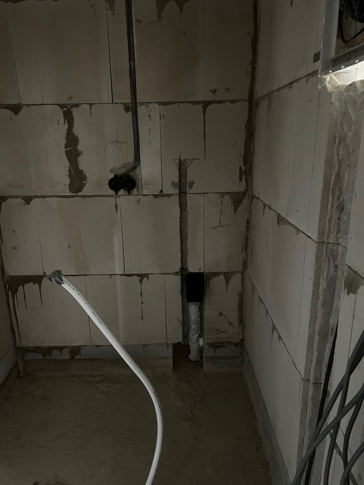 Surowy stan: instalacja wodno-kanalizacyjna w trakcie montażu w pomieszczeniu z bloczków betonowych. Widoczne rury, kable i przygotowane otwory w ścianach.