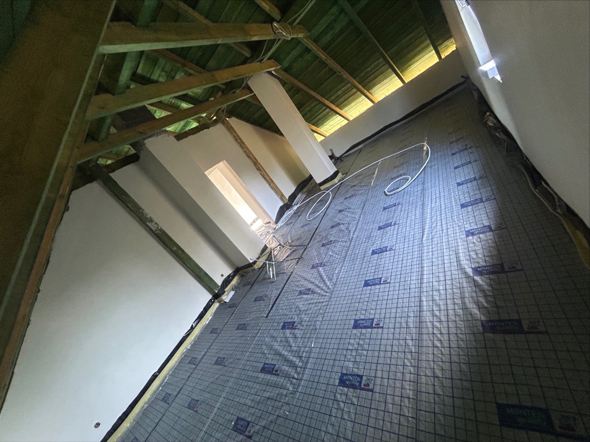 Skośny kadr na poddasze w trakcie izolacji. Widoczne drewniane elementy konstrukcyjne, okno dachowe i folia izolacyjna z nadrukiem siatki i logo 'MONTER Quality'.