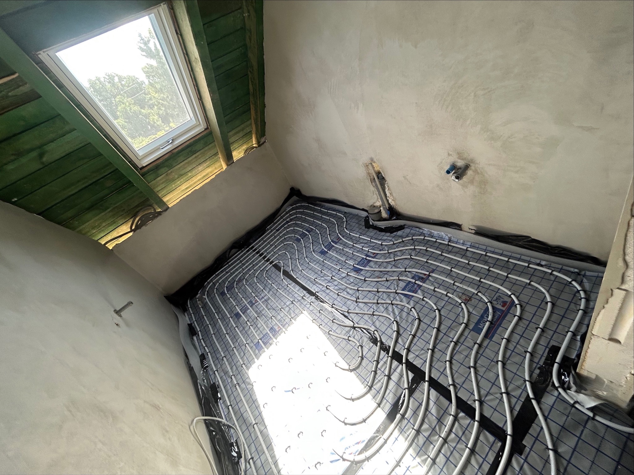 Białe rury ogrzewania podłogowego ułożone na szarej macie w surowym wnętrzu z oknem dachowym i widocznymi elementami instalacji na ścianie.