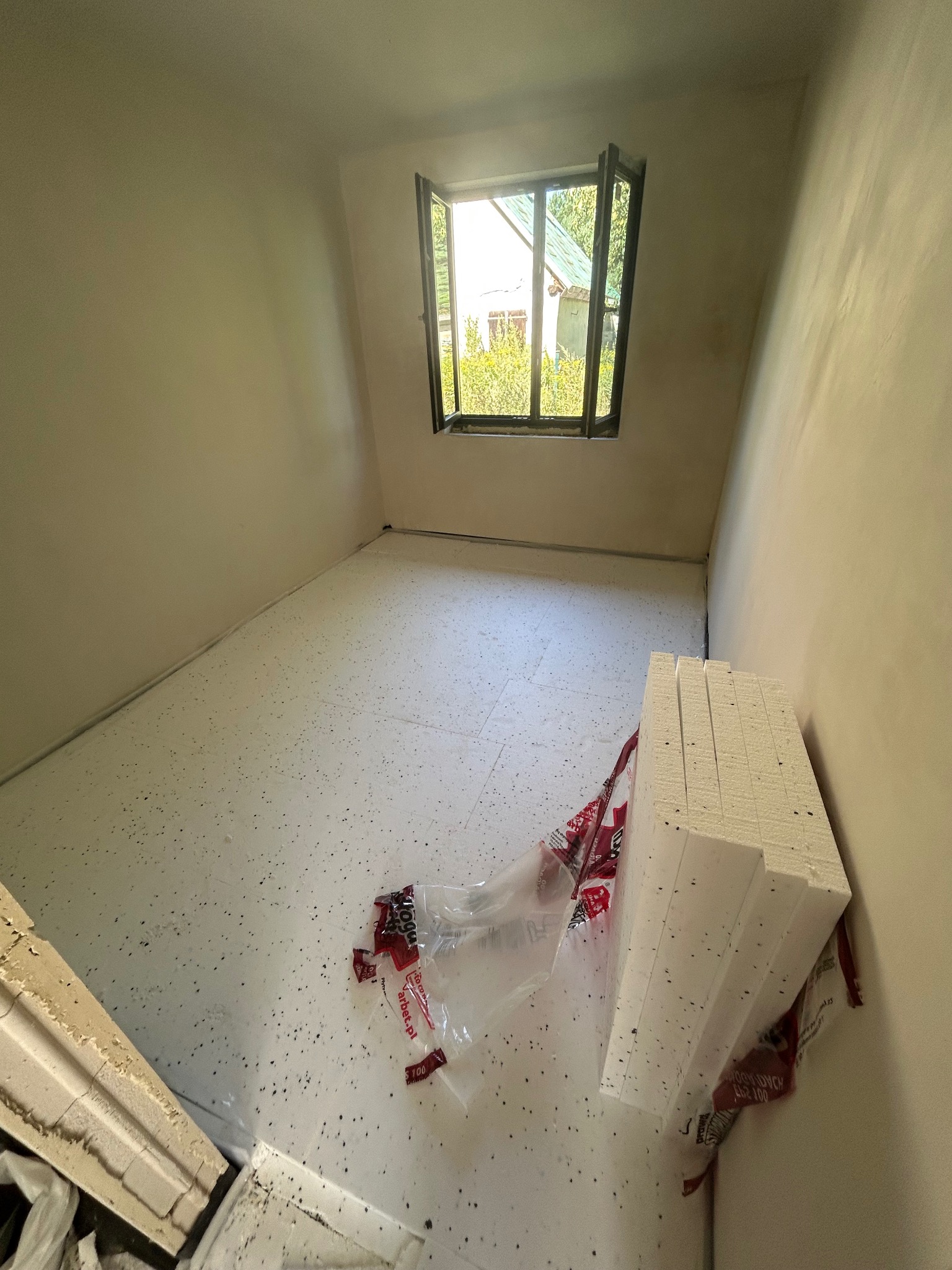 Przygotowanie do montażu ogrzewania podłogowego: rozłożony styropian z folią w pustym pomieszczeniu z oknem. Widoczne płyty styropianowe i fragment ściany.