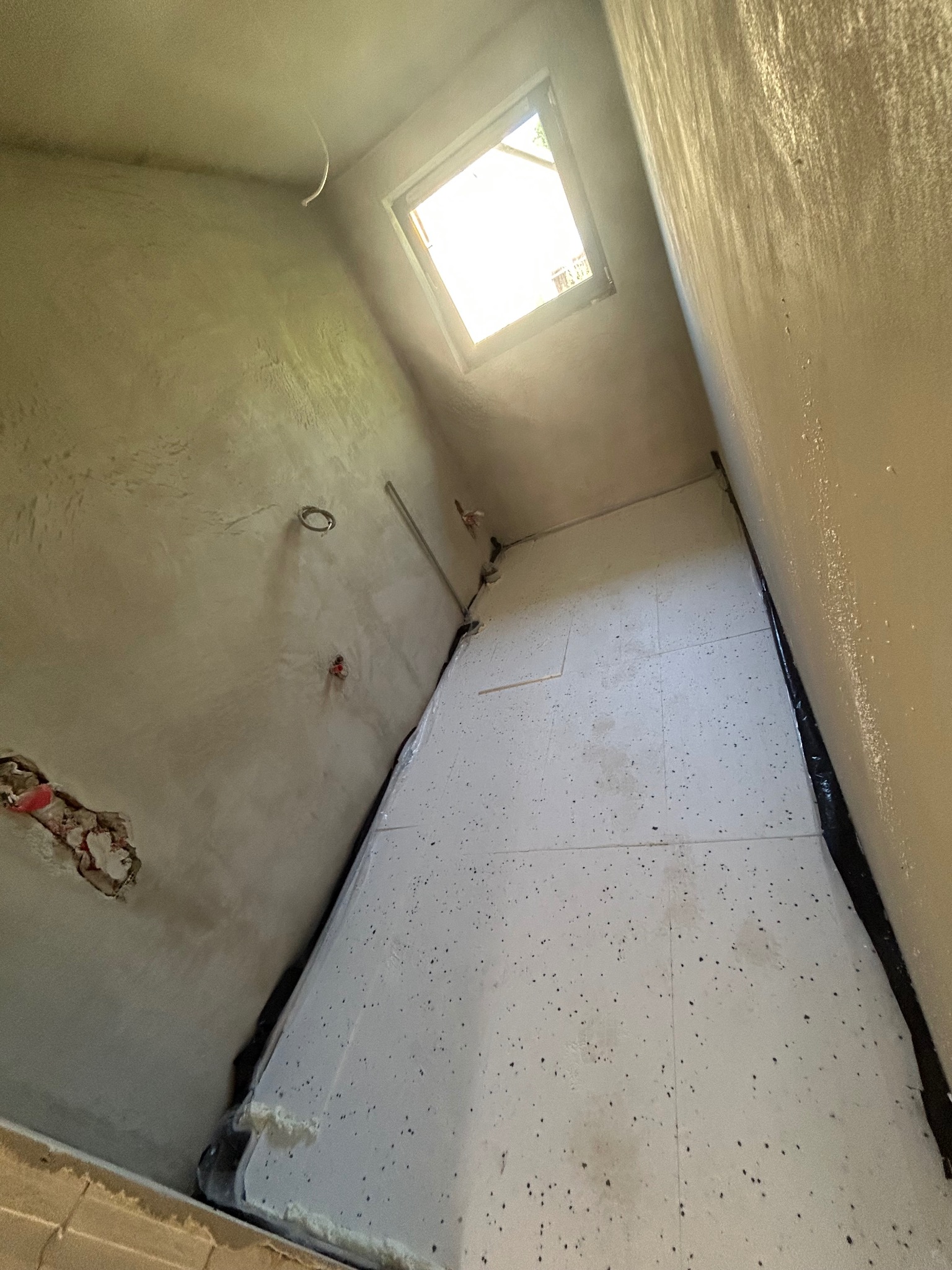 Białe płyty izolacyjne pod ogrzewanie podłogowe ułożone w surowym pomieszczeniu z oknem dachowym. Widoczne przewody instalacji elektrycznej i fragment ściany z uszkodzonym tynkiem.