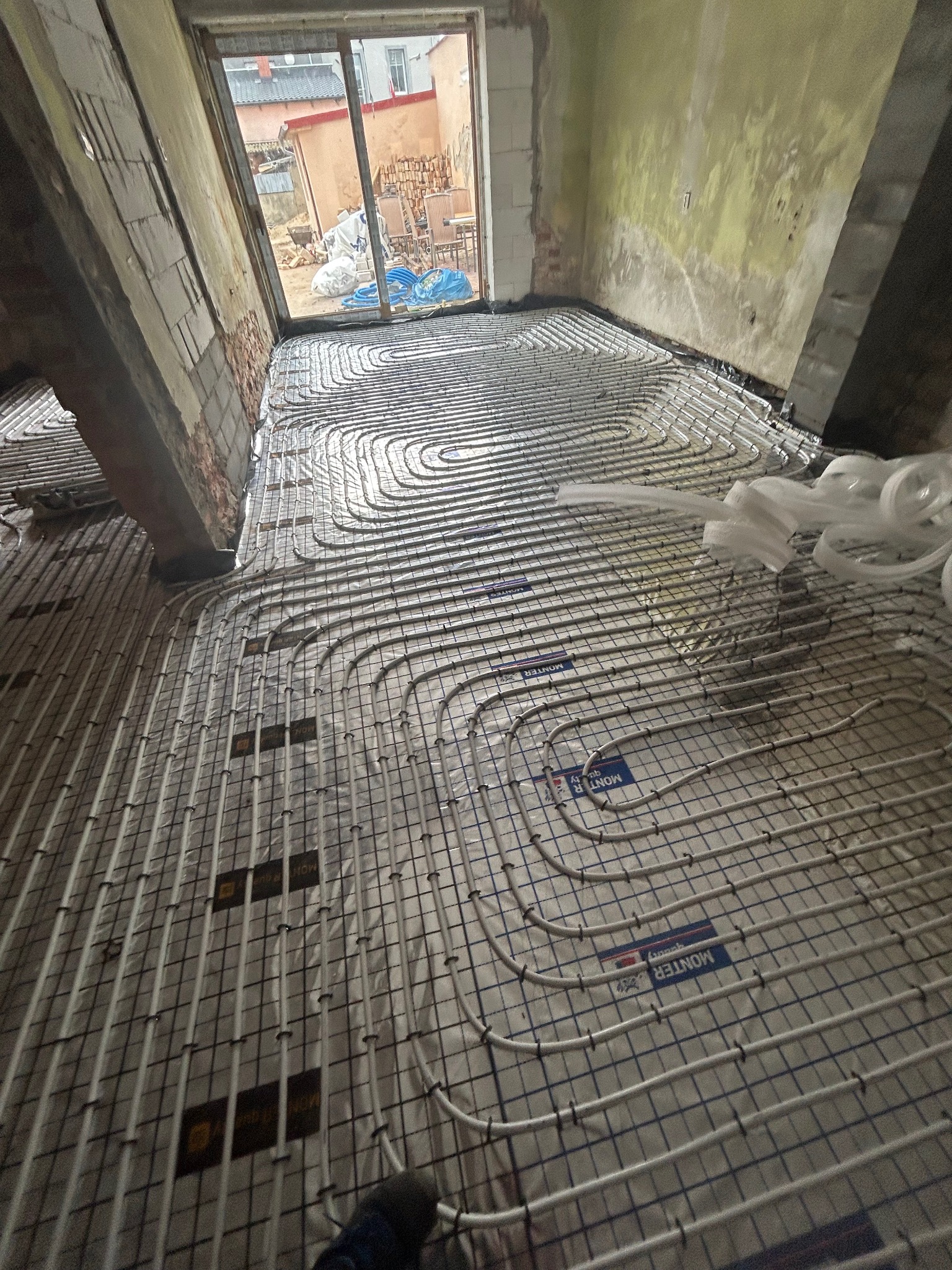 Ułożone rury ogrzewania podłogowego na metalowej siatce w surowym wnętrzu. Widoczne fragmenty ścian w trakcie remontu i okno z widokiem na zewnątrz.