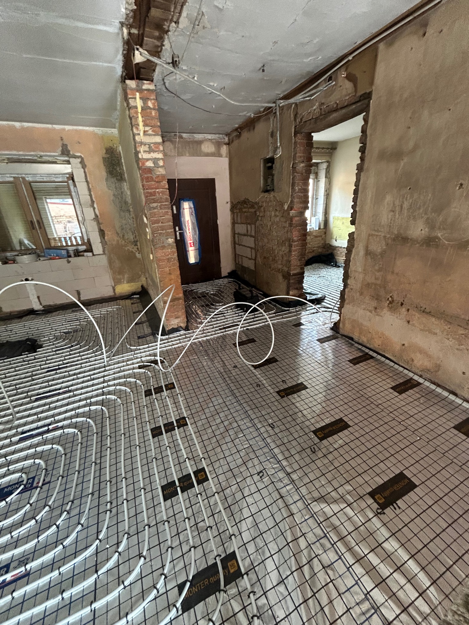 Ułożone białe rury ogrzewania podłogowego na srebrnej folii z czarną siatką w surowym wnętrzu remontowanego mieszkania z widocznymi cegłami i instalacjami.