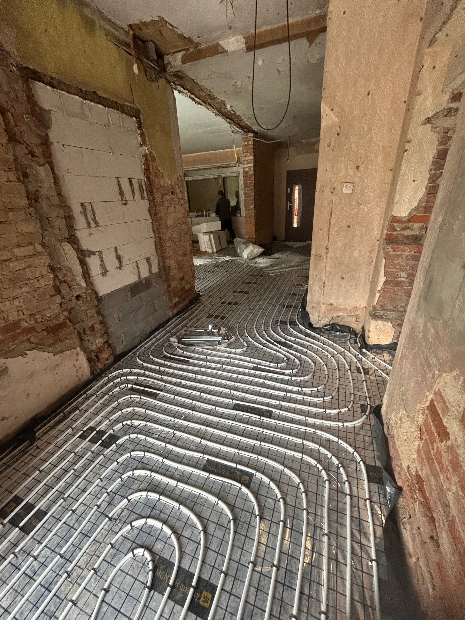Białe rury ogrzewania podłogowego ułożone na czarnej folii w remontowanym wnętrzu z odsłoniętą cegłą i widoczną instalacją elektryczną.