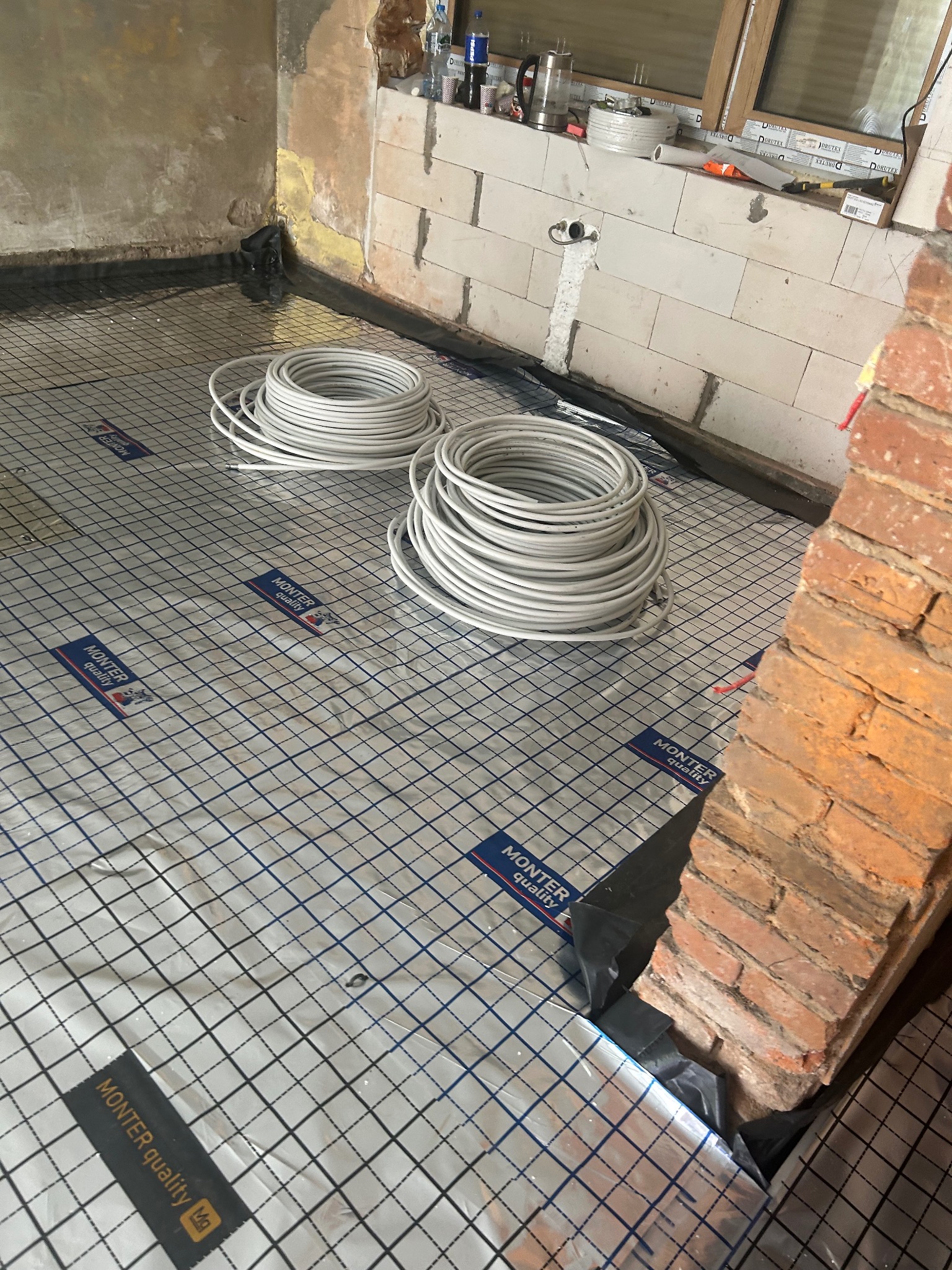 Rozłożona mata grzewcza z przewodami w zwojach, przygotowana do montażu ogrzewania podłogowego. Widoczne materiały izolacyjne i fragment ściany z cegły.