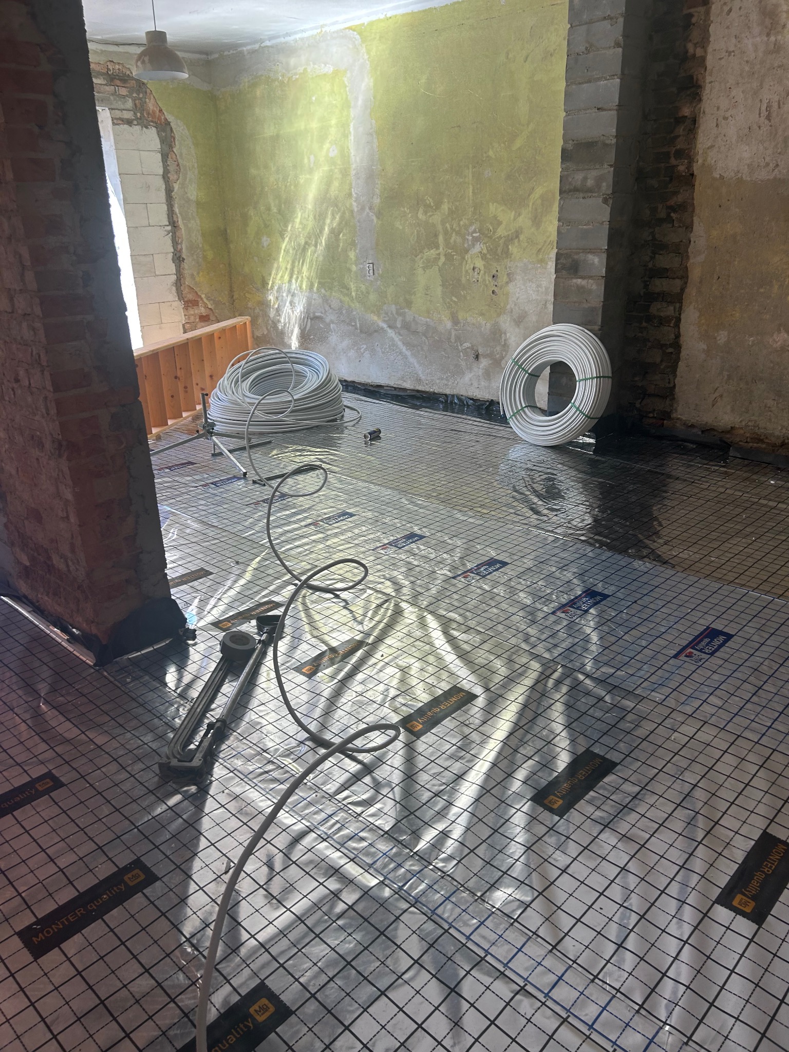 Przygotowana podłoga pod ogrzewanie podłogowe z rozłożoną folią izolacyjną i rurami zwiniętymi w kręgi. Widoczne narzędzia i surowe ściany w tle.