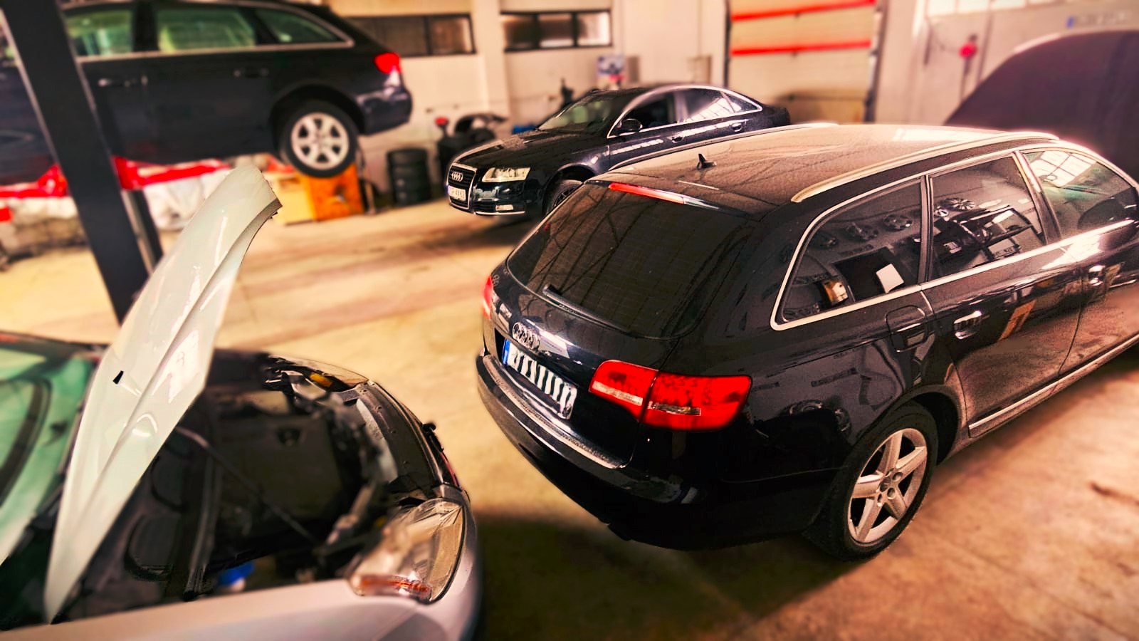 Warsztat samochodowy: kilka aut marki Audi, w tym jeden na podnośniku, z otwartą maską, w trakcie naprawy. Widoczne narzędzia i wyposażenie warsztatu.