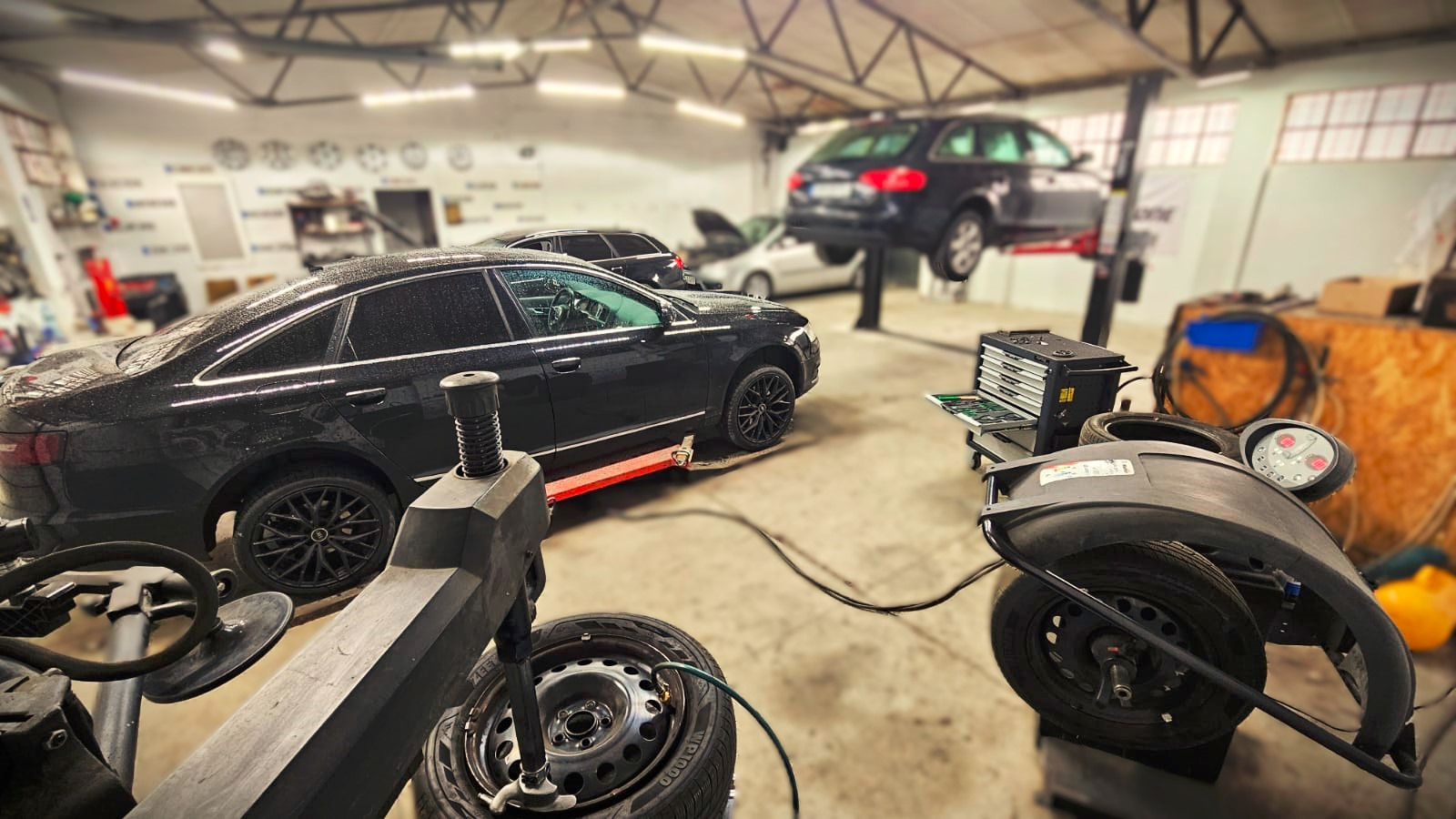 Warsztat samochodowy: czarny sedan na podnośniku, wyważarka kół na pierwszym planie, inne auta w tle. Widoczne narzędzia i wyposażenie warsztatu.