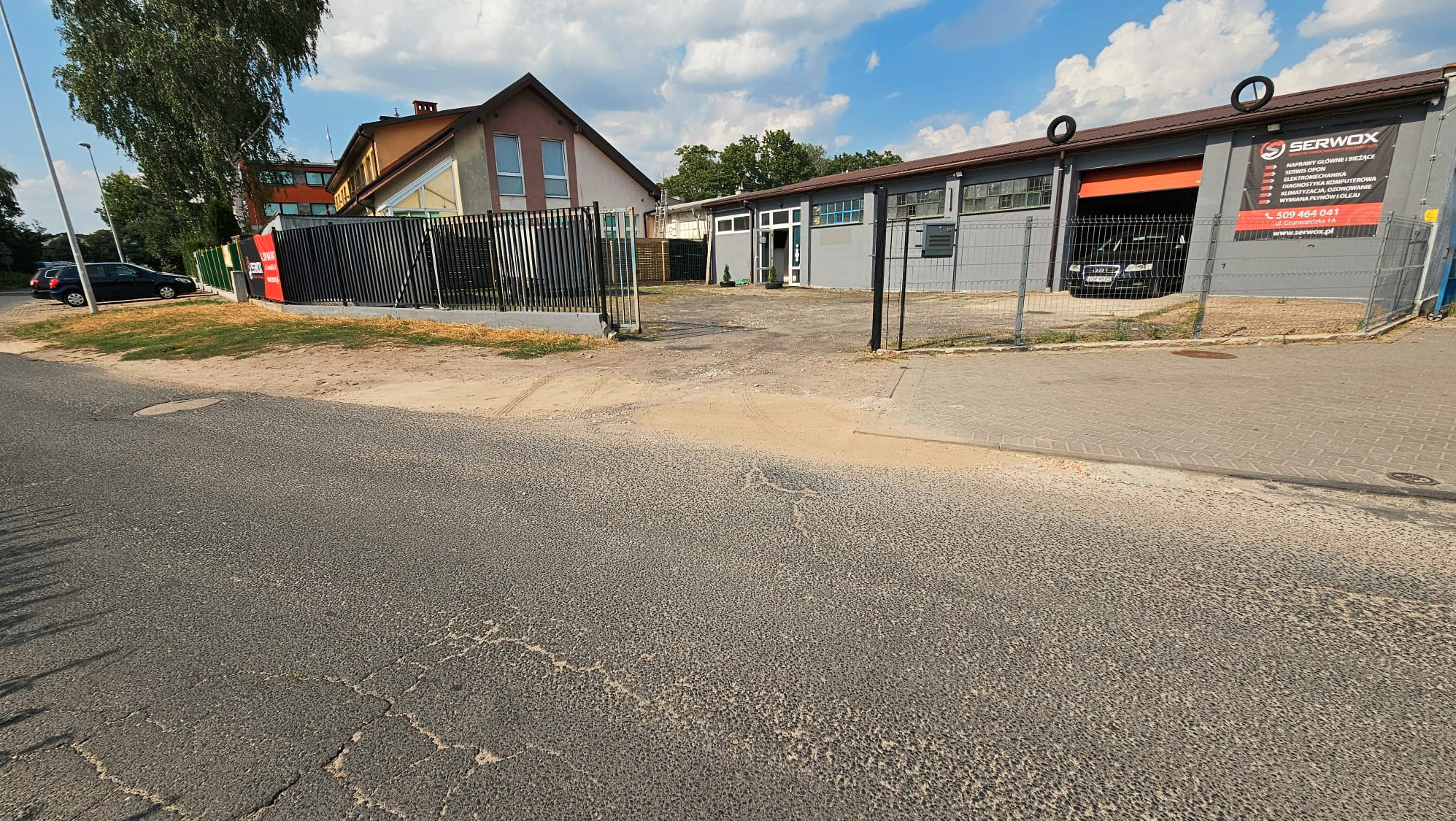 Widok na wjazd do warsztatu samochodowego Serwox z otwartą bramą, za którą stoi czarny samochód. Na budynku reklama z zakresem usług i numerem telefonu.