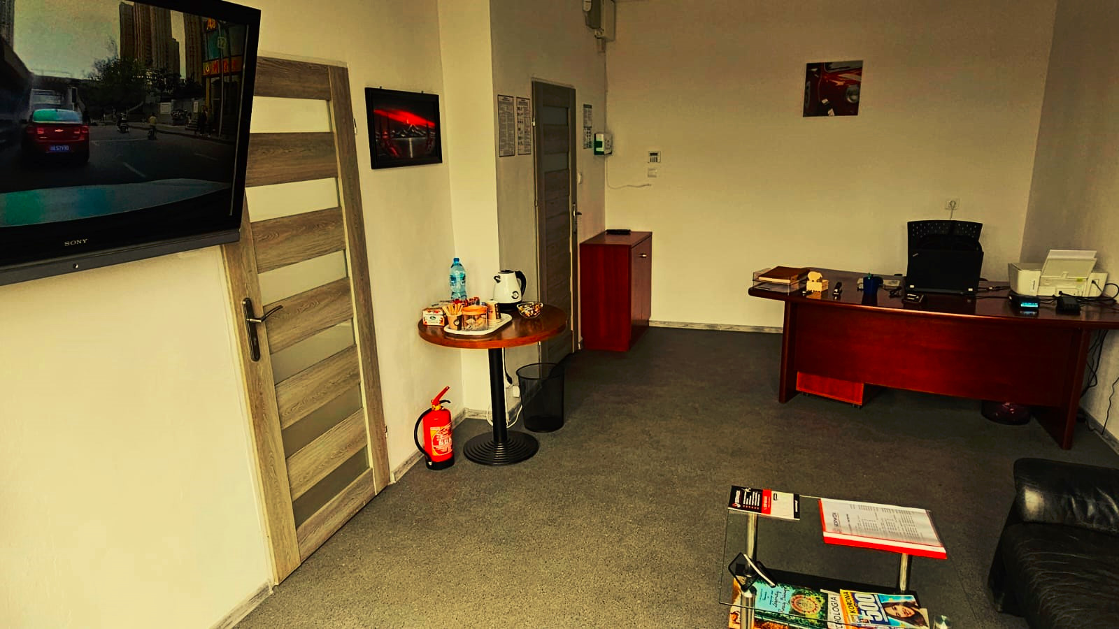 Wnętrze biura warsztatu samochodowego: biurko z laptopem, stolik z poczęstunkiem, gaśnica, telewizor z widokiem z kamery samochodowej. Umeblowane, jasne pomieszczenie.