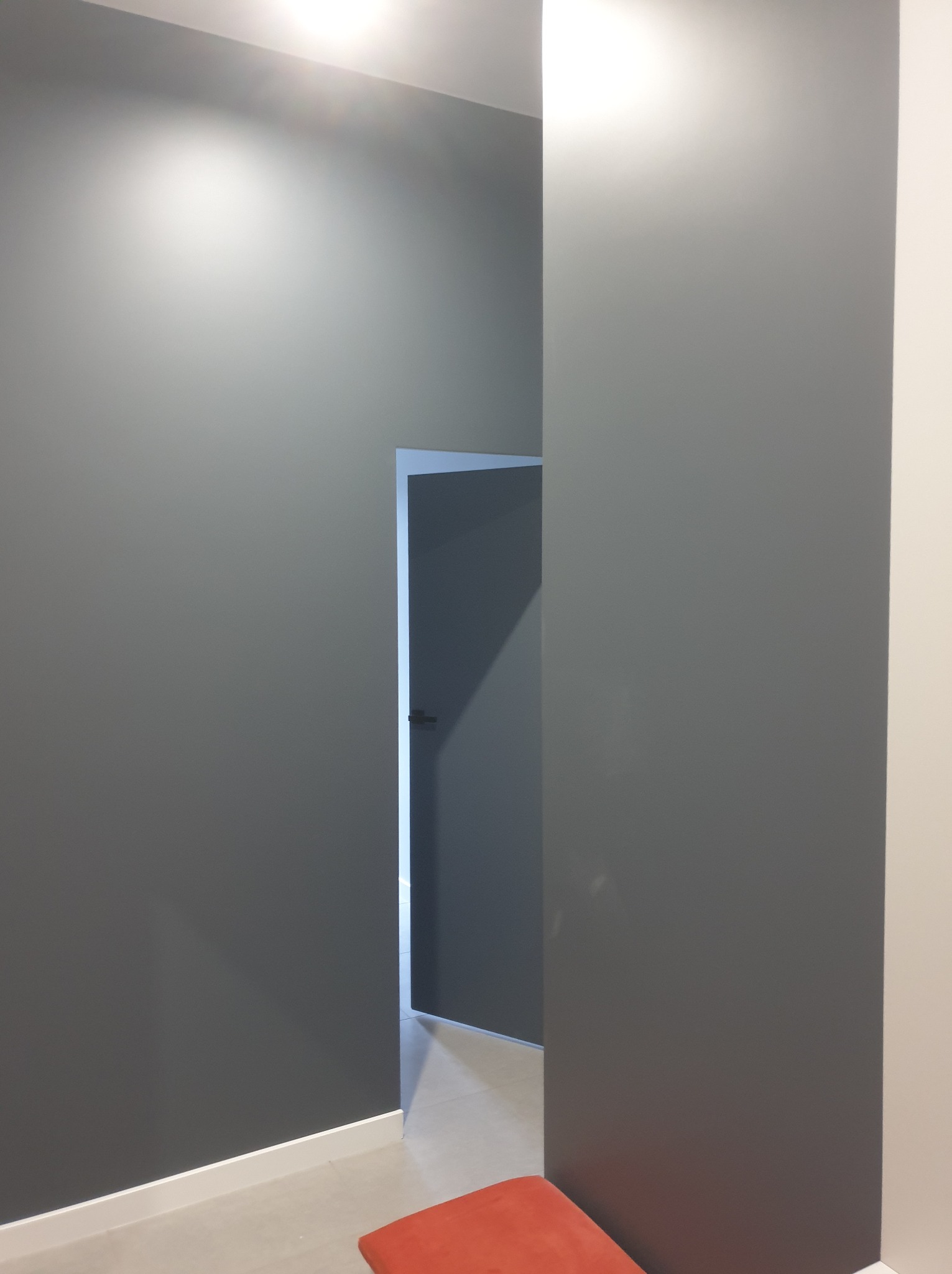 Szara ściana po malowaniu z otwartymi drzwiami i fragmentem czerwonego siedziska. Minimalistyczne wykończenie wnętrza z listwami przypodłogowymi.