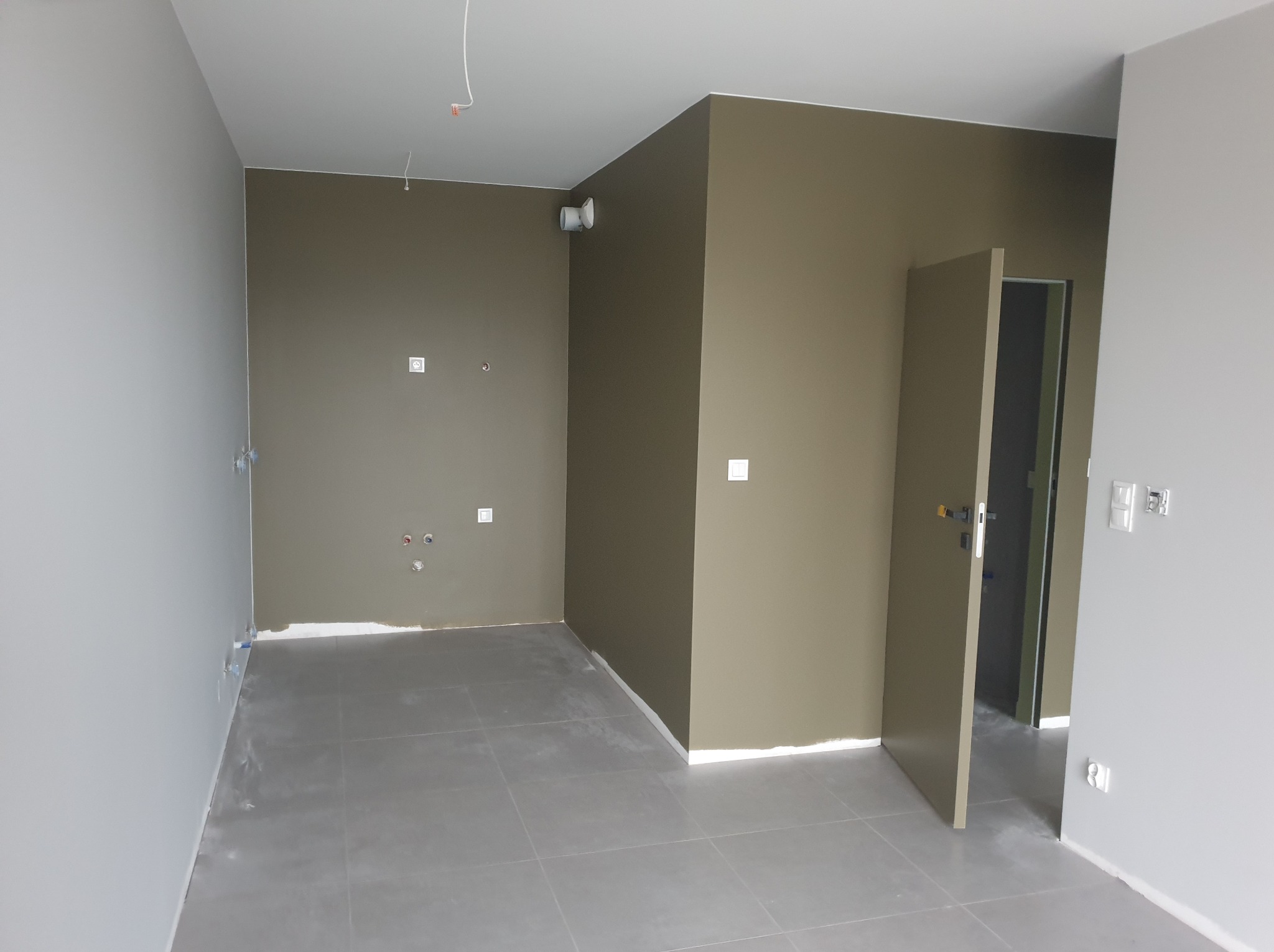 Pokój po malowaniu ścian na dwa kolory: szary i oliwkowy. Widoczne gniazdka elektryczne i otwarta, oliwkowa  futryna drzwiowa. Szare płytki na podłodze.